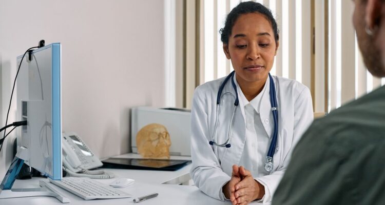 A doctor in a white coat listening to a patient across a desk in a consultation room