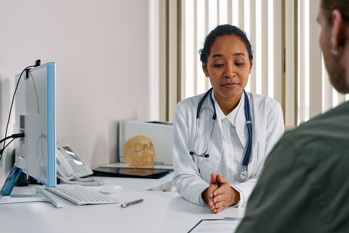 A doctor in a white coat listening to a patient across a desk in a consultation room