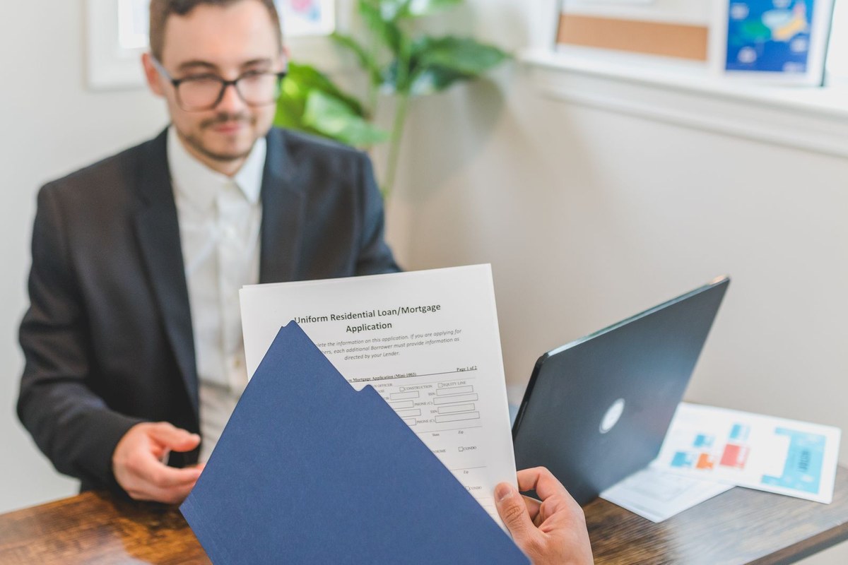 Hands passing a blue folder with a loan application across a desk to a man in a blazer and glasses