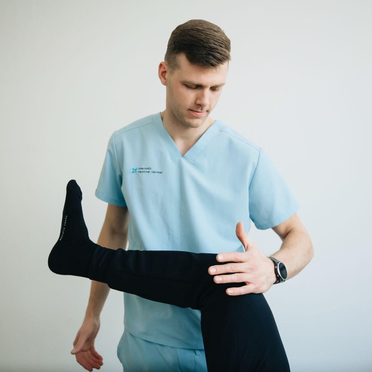 A clinician in light blue scrubs supporting a patient's raised, bent leg to check knee and hip movement